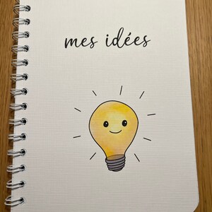 Pu&ograve; includere: Un quaderno bianco con rilegatura a spirale. La copertina ha il testo "mes id&eacute;es" e una lampadina gialla con un viso sorridente.