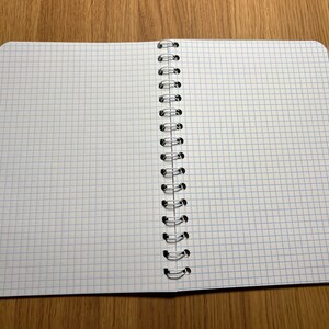 Pu&ograve; includere: Un quaderno a spirale aperto con pagine bianche e linee di griglia blu. Il quaderno &egrave; disteso su una superficie di legno.