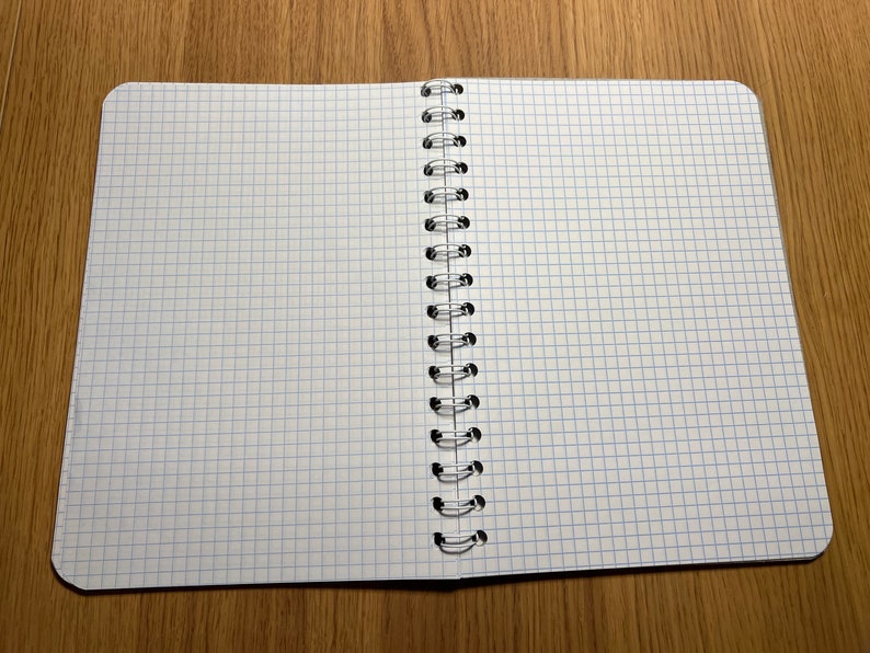 Pu&ograve; includere: Un quaderno a spirale aperto con pagine bianche e linee di griglia blu. Il quaderno &egrave; disteso su una superficie di legno.