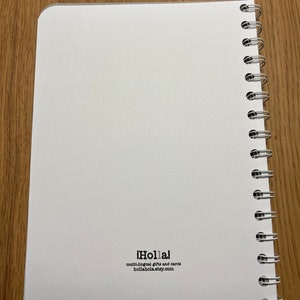 Pu&ograve; includere: Un quaderno bianco a spirale con il testo "&iexcl;Holla!" e "multi-lingual gifts and cards hollahola.etsy.com" stampato sulla copertina posteriore.