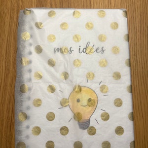 Pu&ograve; includere: Un quaderno bianco con un motivo a pois dorati e il testo "mes id&eacute;es" in nero. Una lampadina gialla con un viso sorridente &egrave; al centro del quaderno.