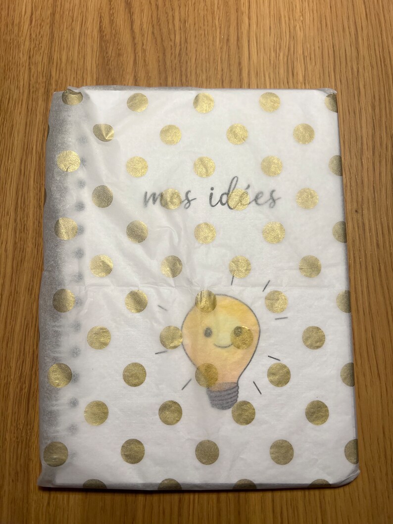 Pu&ograve; includere: Un quaderno bianco con un motivo a pois dorati e il testo "mes id&eacute;es" in nero. Una lampadina gialla con un viso sorridente &egrave; al centro del quaderno.