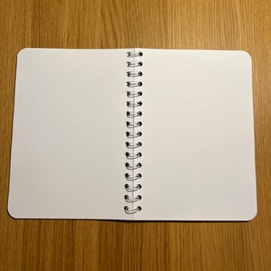 Pu&ograve; includere: Un quaderno bianco a spirale con pagine vuote, aperto e piatto su una superficie di legno.