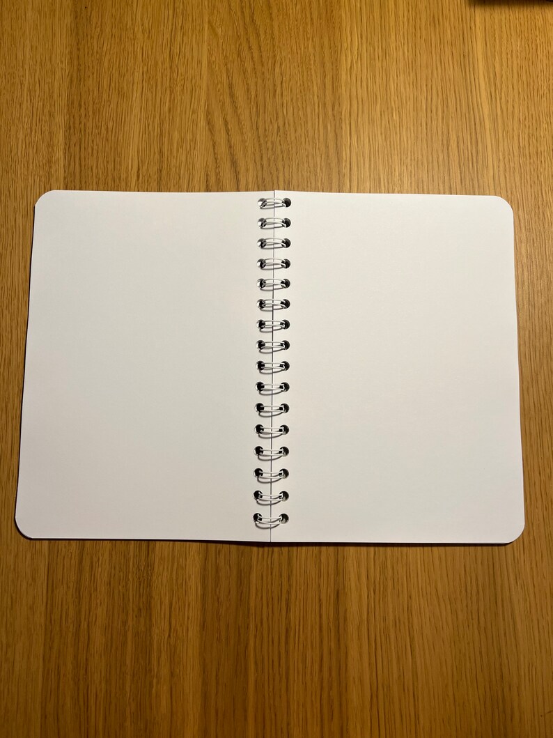 Pu&ograve; includere: Un quaderno bianco a spirale con pagine vuote, aperto e piatto su una superficie di legno.