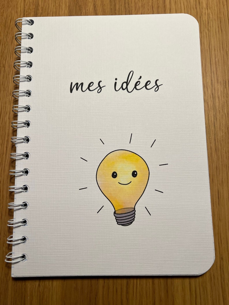 Pu&ograve; includere: Un quaderno bianco con rilegatura a spirale. La copertina ha il testo "mes id&eacute;es" e una lampadina gialla con un viso sorridente.