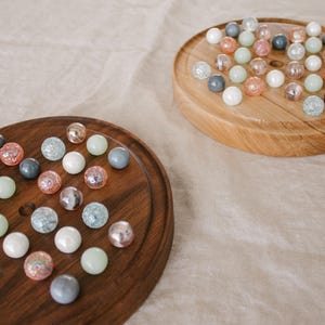 May include: Two wooden game boards with a collection of colorful marbles. The marbles are in various colors, including white, blue, pink, and green. The boards are round and oval, with a smooth, polished finish. The background is a neutral-toned fabric.
