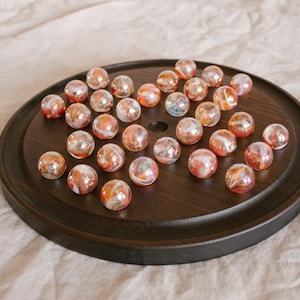 May include: A wooden solitaire board with a dark finish, holding a collection of iridescent marbles. The marbles have a mix of orange, white, and clear colors, creating a shimmering effect. The board is round with a central hole.