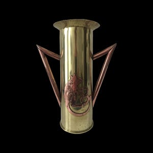 Puede incluir: Un jarrón de latón con detalles de cobre y un diseño decorativo de rata. El jarrón tiene un cuerpo cilíndrico con una parte superior ensanchada y dos asas en forma de V. La rata está representada en una media luna.