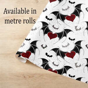 May include: White patterned paper with a repeating design of black bat wings, red hearts, and gray bats. The text "Available in metre rolls" is printed on the paper. The paper is rolled up on a light wood surface.