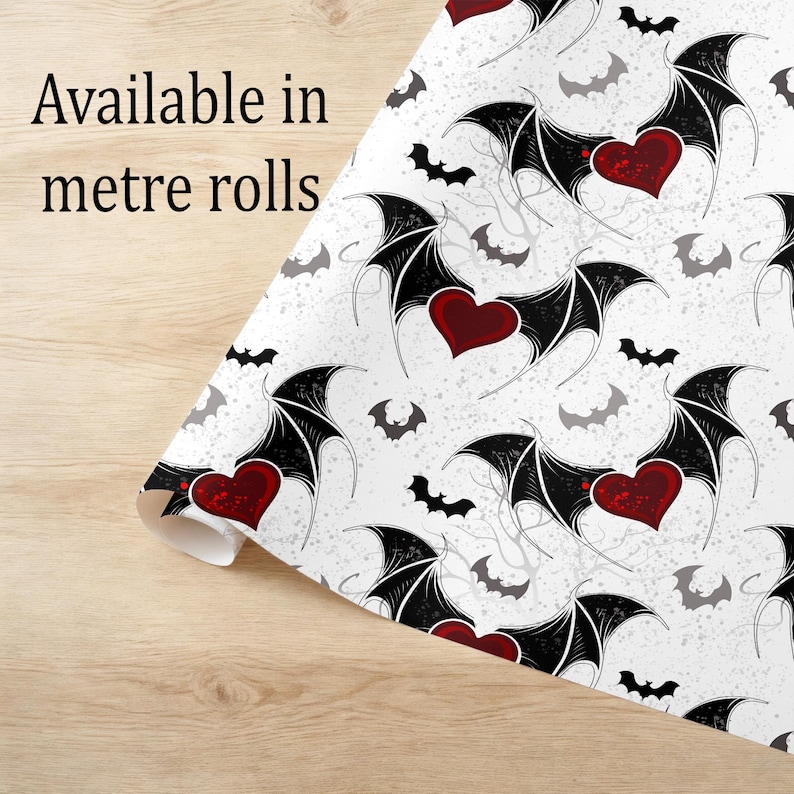 May include: White patterned paper with a repeating design of black bat wings, red hearts, and gray bats. The text "Available in metre rolls" is printed on the paper. The paper is rolled up on a light wood surface.