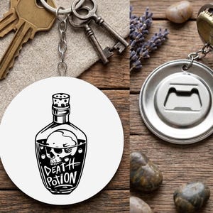 May include: A round white keychain featuring a black illustration of a skull in a bottle, labelled "DEATH POTION." Beside it is a silver bottle opener keychain. The keys are on a wooden surface.
