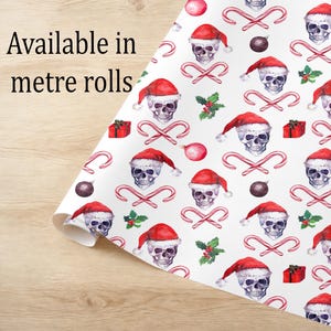 May include: White wrapping paper with a repeating pattern of skulls wearing Santa hats, candy canes, red gift boxes, and ornaments. The text "Available in metre rolls" is visible on the left side of the image.