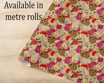 May include: A roll of patterned paper with a repeating design of skulls decorated with pink, red, and cream flowers. The background is a neutral beige colour. The text "Available in metre rolls" is visible.
