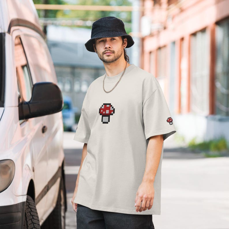 May include: A white t-shirt with a pixelated red and white mushroom graphic on the front. The shirt has a relaxed fit and short sleeves.