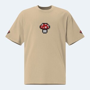 May include: A beige t-shirt with a pixelated red and white mushroom graphic on the front.