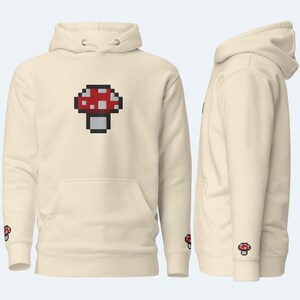 May include: A cream-colored hoodie with a pixelated red and white mushroom graphic on the front. The hoodie has a drawstring hood and a kangaroo pocket.