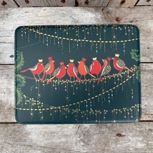 May include: A rectangular, dark teal tin with a decorative design featuring red birds wearing gold crowns perched on a branch. Gold string lights and foliage complete the festive scene.