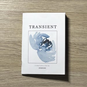 以下が含まれることがあります： 「TRANSIENT」という黒い文字が書かれた白い冊子。表紙には青い水彩画で描かれた花が描かれている。イラストの下には「a short story zine by DEMORE」という文字が書かれている。