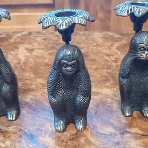 May include: Three antique bronze candle holders, each featuring a monkey figurine in a different pose: covering eyes, covering ears, and covering mouth. The candle holders have a floral design at the top, and are displayed on a wooden surface.