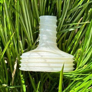 May include: A translucent, off-white bottle stopper with a unique design. The stopper features a ridged neck and a flared, textured base. The object is set against a backdrop of vibrant green grass.
