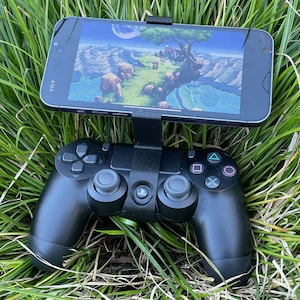 May include: A black game controller with a smartphone attached, displaying a video game scene. The controller rests on a bed of green grass. The smartphone is held in place by a black bracket.