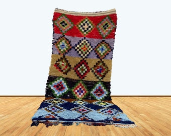 May include: A colorful Moroccan rug with a geometric pattern in shades of red, blue, green, and yellow. The rug is made of wool and has a fringed edge.