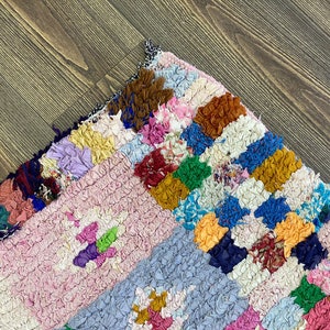 May include: A colorful, handmade rag rug with a patchwork design. The rug features a variety of colors, including pink, blue, yellow, and brown. The texture is thick and textured, with a mix of fabric scraps. The rug is placed on a wooden floor.