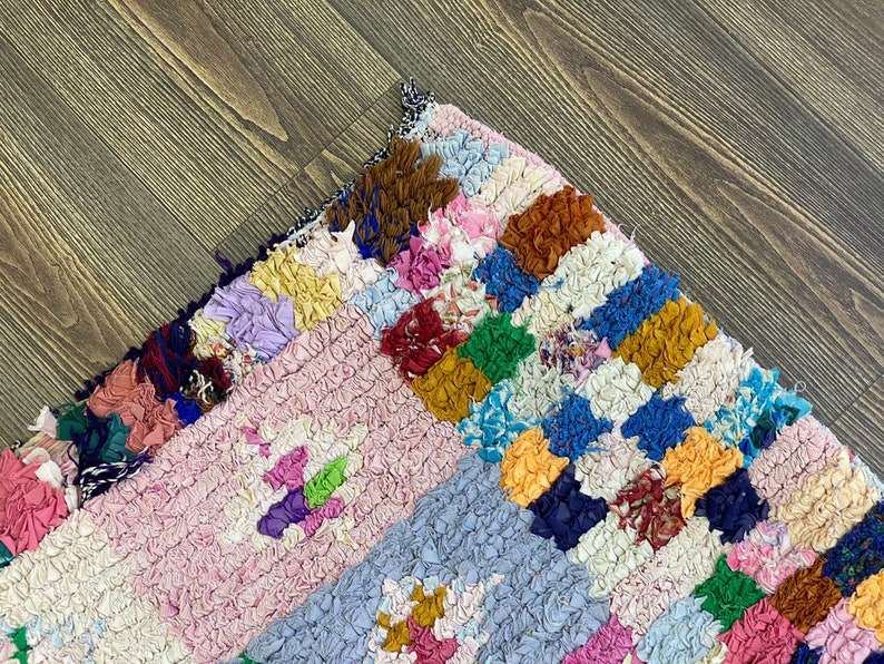 May include: A colorful, handmade rag rug with a patchwork design. The rug features a variety of colors, including pink, blue, yellow, and brown. The texture is thick and textured, with a mix of fabric scraps. The rug is placed on a wooden floor.