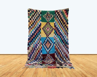 May include: A colorful Moroccan rug with a geometric pattern in shades of blue, green, yellow, red, and pink. The rug has a diamond-shaped design in the center and is woven with a thick, textured pile.