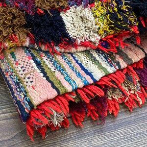 Pu&ograve; includere: Un tappeto tessuto colorato con un bordo a frangia. Il tappeto &egrave; fatto di filati di diversi colori e ha un design a patchwork. Il tappeto &egrave; piegato a met&agrave; e la frangia &egrave; visibile.