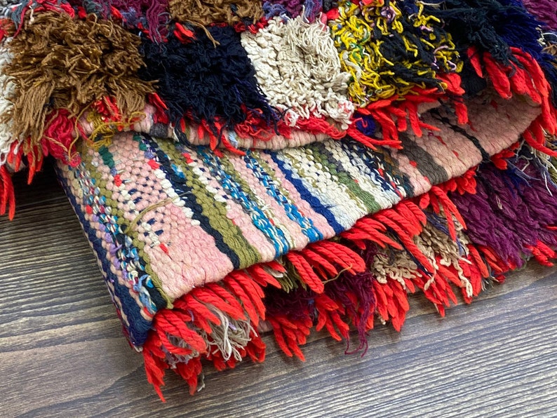 Pu&ograve; includere: Un tappeto tessuto colorato con un bordo a frangia. Il tappeto &egrave; fatto di filati di diversi colori e ha un design a patchwork. Il tappeto &egrave; piegato a met&agrave; e la frangia &egrave; visibile.