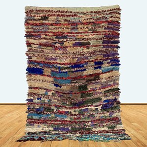 May include: A rectangular, multi-colored rug with a textured, woven appearance. The rug features horizontal stripes in shades of beige, red, blue, purple, and green. The rug is displayed against a white background and a wooden floor.