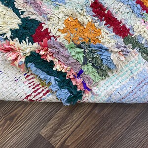 Puede incluir: Una colorida alfombra de trapo hecha a mano con retazos de tela en tonos azules, verdes, rojos y naranjas. La alfombra tiene un aspecto tejido y texturizado y est&aacute; parcialmente enrollada, revelando la parte inferior.