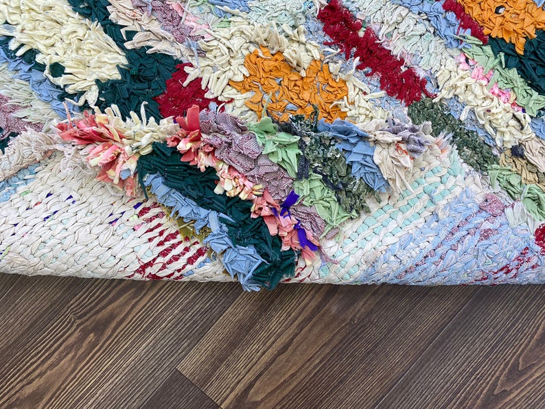 Puede incluir: Una colorida alfombra de trapo hecha a mano con retazos de tela en tonos azules, verdes, rojos y naranjas. La alfombra tiene un aspecto tejido y texturizado y est&aacute; parcialmente enrollada, revelando la parte inferior.