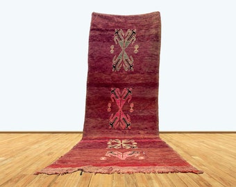 May include: A long, rectangular, hand-woven rug in shades of red and pink. The rug features three symmetrical geometric designs in black, white, and green. The rug is displayed on a wooden floor.
