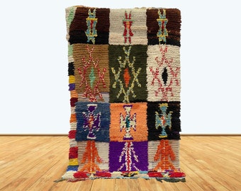 May include: A colorful, rectangular rug with a patchwork design. The rug features a variety of colors, including brown, green, orange, purple, and blue. Each square has a unique pattern, with geometric and abstract designs. The rug is displayed against a white background.