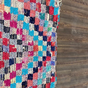 May include: A colorful, handwoven rug with a checkered pattern in shades of pink, red, blue, white, and black. The rug is made of recycled materials and has a unique, textured surface.