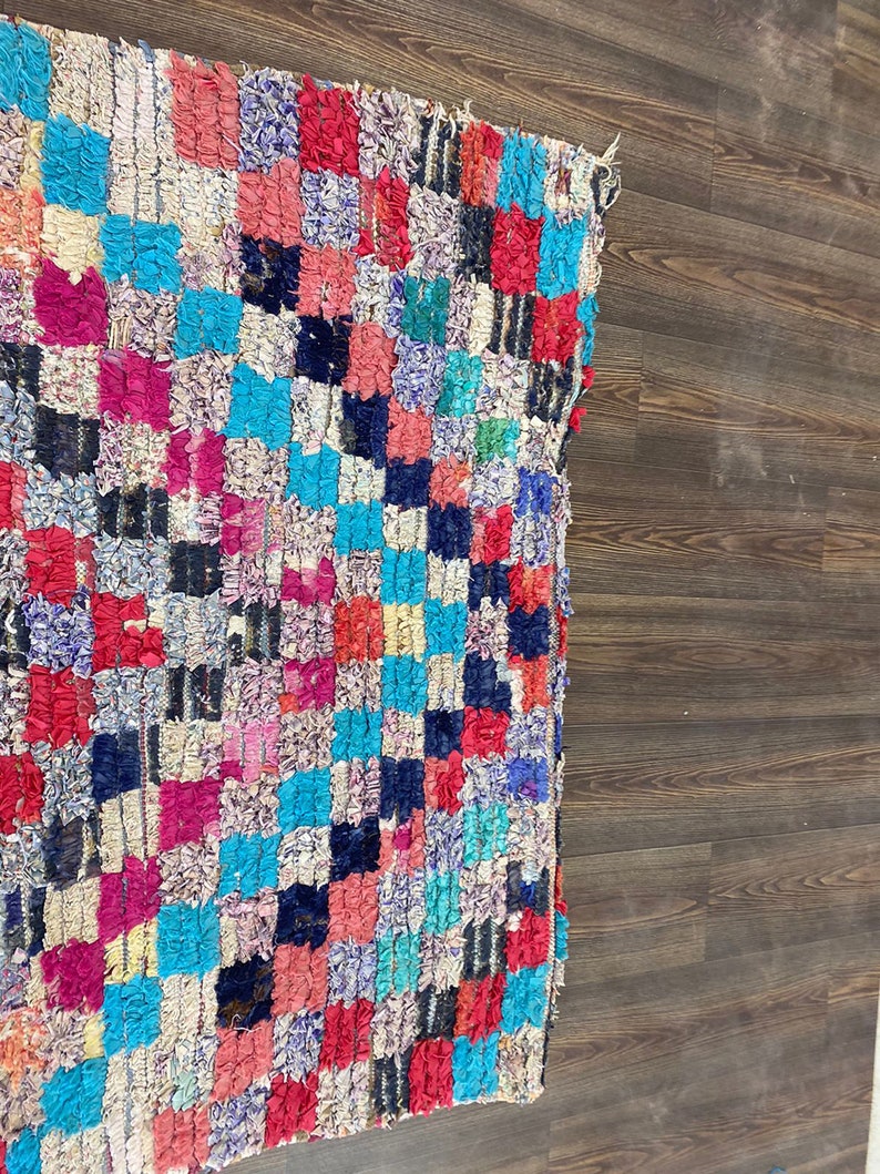 May include: A colorful, handwoven rug with a checkered pattern in shades of pink, red, blue, white, and black. The rug is made of recycled materials and has a unique, textured surface.