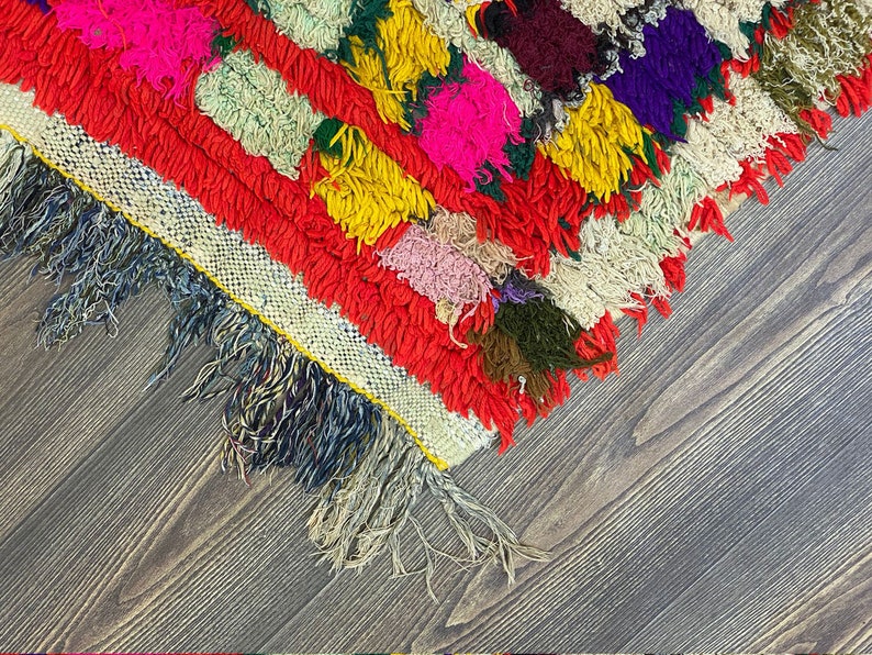 Pu&ograve; includere: Un tappeto colorato tessuto a mano con un bordo a frangia. Il tappeto &egrave; fatto di lana e ha una variet&agrave; di colori, tra cui rosso, giallo, verde, blu e rosa.