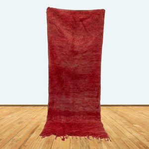 May include: A long, red Moroccan rug with fringe on the bottom. The rug is hanging on a white wall with a wooden floor underneath.
