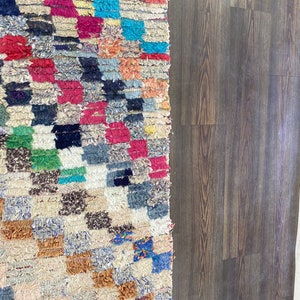 May include: A colorful Moroccan rug with a geometric pattern in shades of red, blue, green, and brown. The rug is made of woven wool and has a fringed edge.