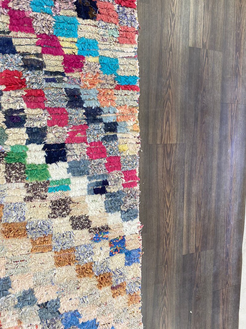 May include: A colorful Moroccan rug with a geometric pattern in shades of red, blue, green, and brown. The rug is made of woven wool and has a fringed edge.
