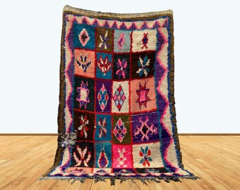 May include: A colorful, rectangular Moroccan rug with a geometric pattern. The rug features a grid of squares in various colors, including blue, pink, purple, and brown. The edges are trimmed with a dark brown border and fringe.