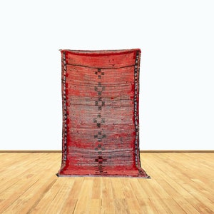 Puede incluir: Una alfombra tejida roja y negra con un patrón geométrico. La alfombra está colgada en una pared blanca y se encuentra en una habitación con suelo de madera.