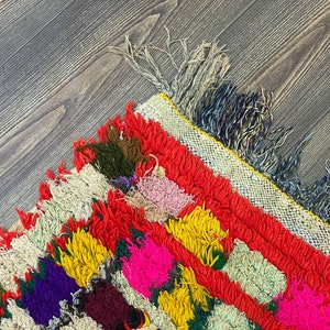 Pu&ograve; includere: Un tappeto marocchino colorato con un bordo a frangia. Il tappeto &egrave; fatto di lana e ha una variet&agrave; di colori, tra cui rosso, giallo, verde e viola.