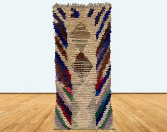 Puede incluir: Una alfombra larga y estrecha, tejida a mano, con un patrón geométrico en tonos de azul, morado, rojo, verde y marrón. La alfombra tiene un fondo blanco y flecos en la parte superior e inferior.