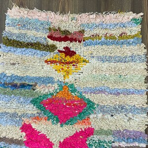Puede incluir: Una colorida alfombra de trapo hecha a mano con una superficie texturizada. La alfombra presenta una vibrante mezcla de colores, incluyendo rosa, azul, verde, amarillo y blanco. El dise&ntilde;o incluye patrones de diamantes y rayas.