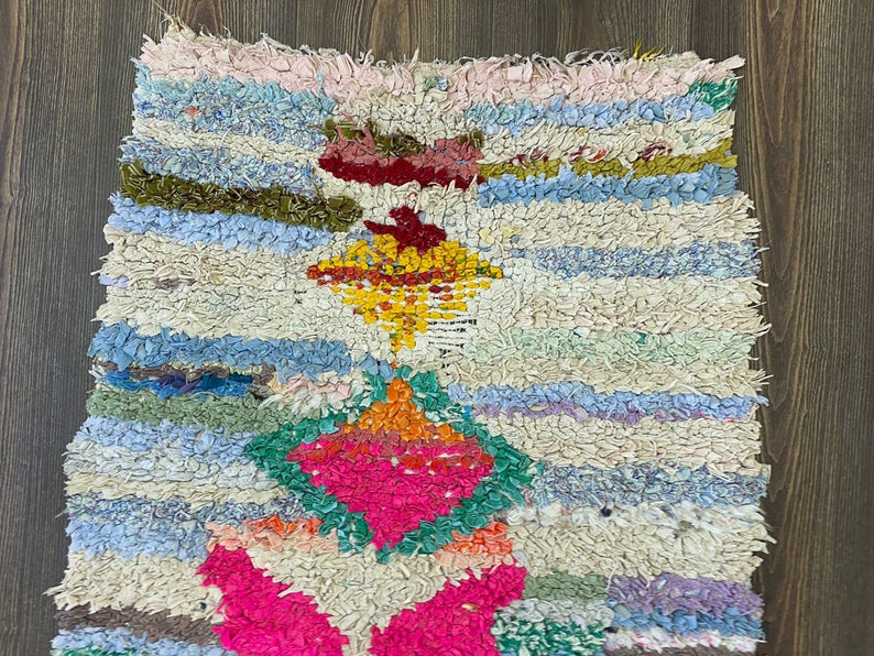 Puede incluir: Una colorida alfombra de trapo hecha a mano con una superficie texturizada. La alfombra presenta una vibrante mezcla de colores, incluyendo rosa, azul, verde, amarillo y blanco. El dise&ntilde;o incluye patrones de diamantes y rayas.