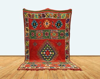 May include: A vibrant red rug with intricate geometric patterns in various colors. The rug features a central diamond design and borders with floral and geometric motifs. The rug is displayed on a wooden floor.