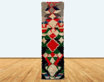 Puede incluir: Una alfombra larga y estrecha con un patrón geométrico en tonos de rojo, verde, negro y blanco. La alfombra está hecha de un material suave y esponjoso y tiene un borde con flecos.